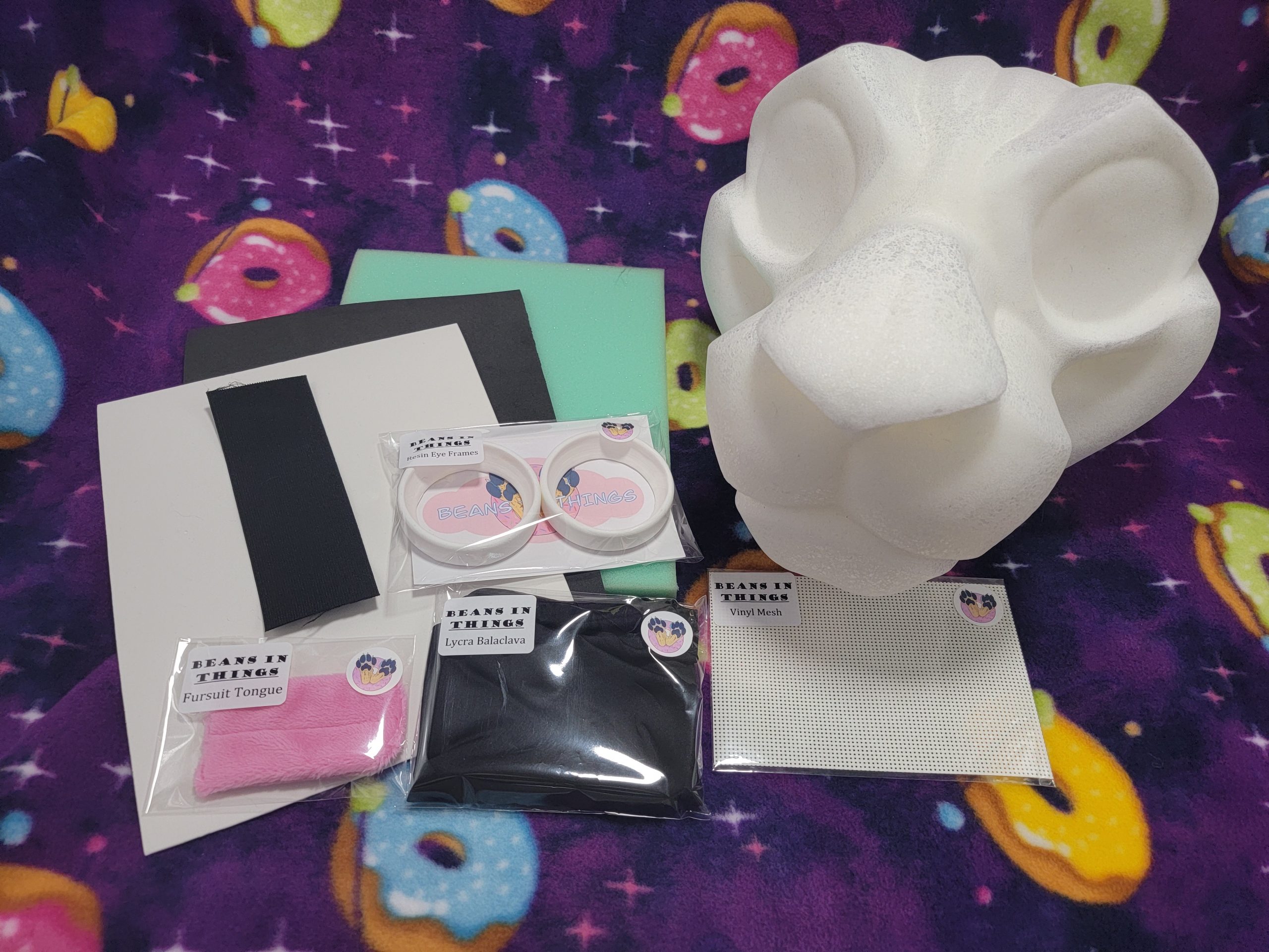 A kit for creating your own big cat costume is shown. To the right is a large cat base made from soft foam, surrounded by foam sheets, resin eye frames, a pink fabric tongue, a pouch with a folded balaclava and two strips of elastic.