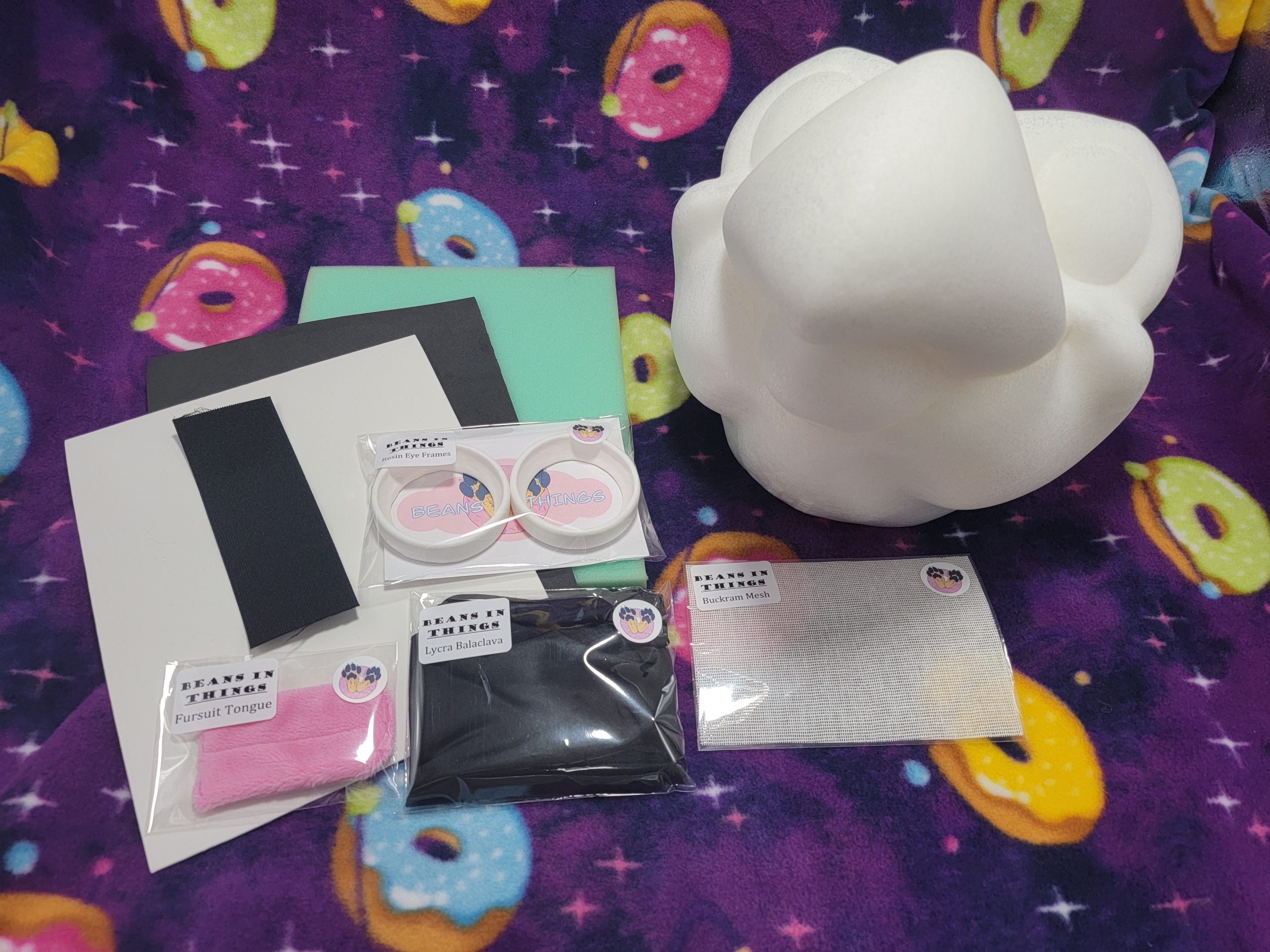 A kit for creating your own bird costume is shown. To the right is a cartoon bird base made from soft foam, surrounded by foam sheets, resin eye frames, a pink fabric tongue, a pouch with a folded balaclava and two strips of elastic.