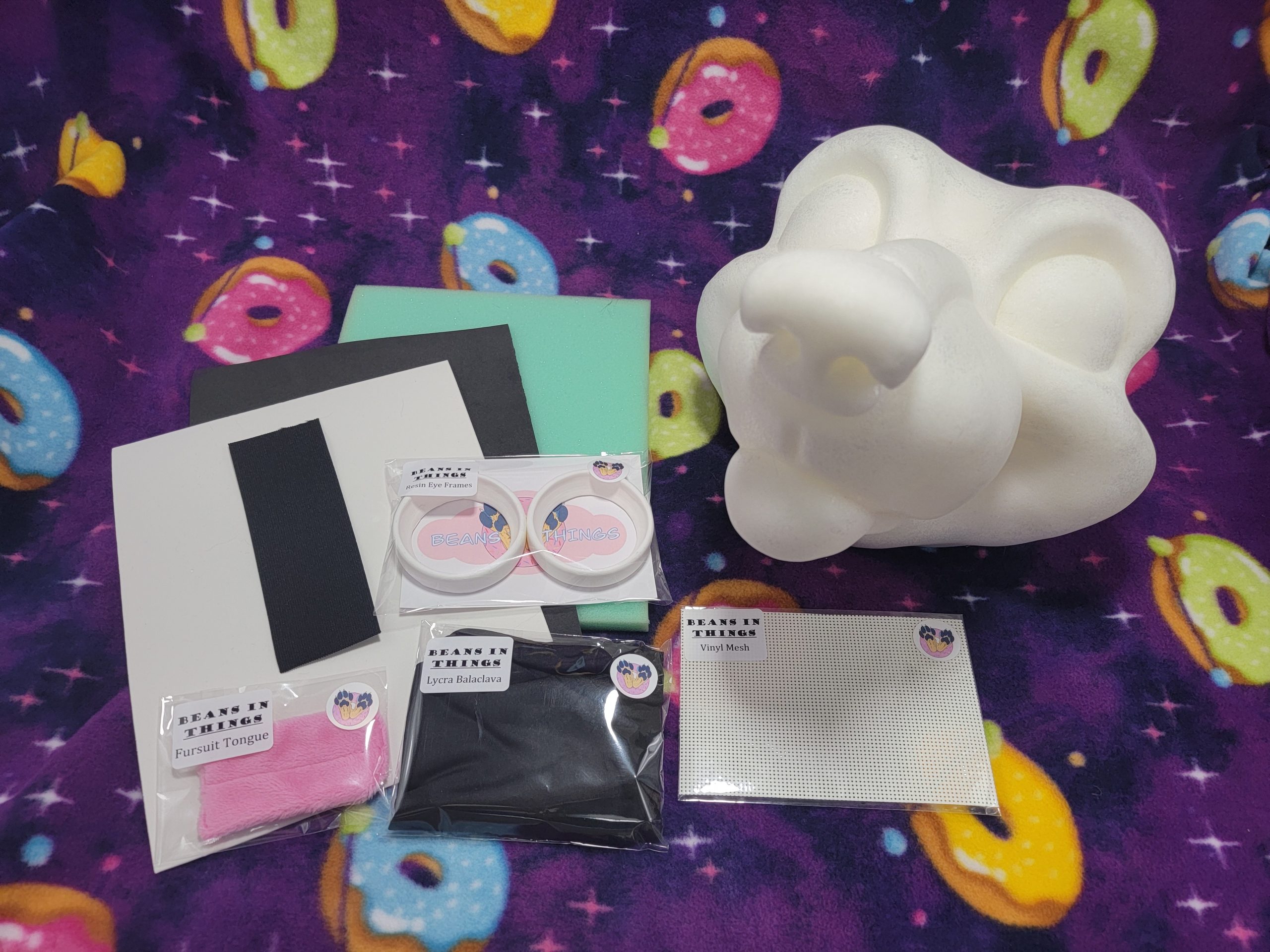 A kit for creating your own wolf costume is shown. To the right is a cartoon wolf base made from soft foam, surrounded by foam sheets, resin eye frames, a pink fabric tongue, a pouch with a folded balaclava and two strips of elastic.